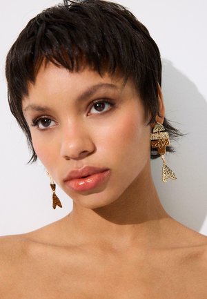 Mlada osoba s kratkom tamnom kosom nosi teksturirane zlatne geometrijske viseće naušnice, gleda blago prema gore na bijeloj pozadini.