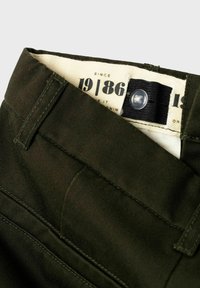 Sötétzöld pamut nadrág, amelyen jól látható derékpánt található gombbal és címkével. Az anyag enyhe textúrával és varrási részletekkel rendelkezik.