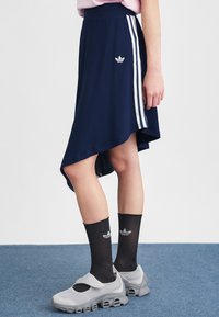 Persona, kas tērpusies tumši zila asimetriskā Adidas svārkā ar baltajām svītrām, melnās Adidas zeķēs un pelēkos acs tīklus apavos ar biezām zolēm.