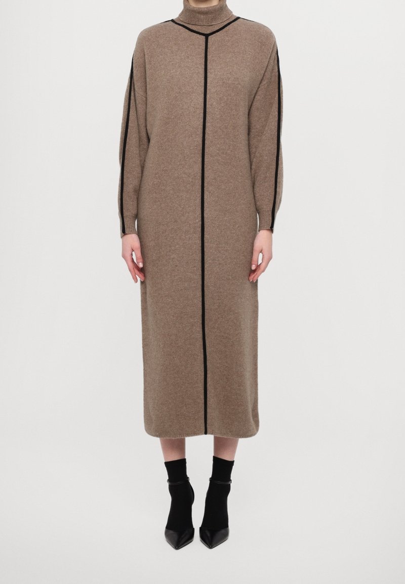 Femme portant une longue robe marron col roulé avec des rayures noires verticales et sur les épaules, associée à des bottines noires sur fond blanc.