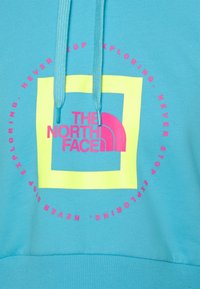 Sweat à capuche bleu clair avec un carré jaune et un texte rose "The North Face" et une phrase circulaire "Never stop exploring" autour du motif.