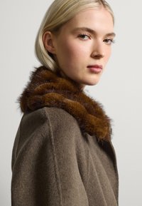 Cappotto di lana marrone con un morbido colletto di pelliccia marrone, texture liscia e design strutturato, visto di lato su uno sfondo neutro.