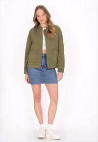 Olivengrön vadderad bomberjacka med två framfickor, bärs över en vit crop top och en denim mini kjol, ihop med vita sneakers.