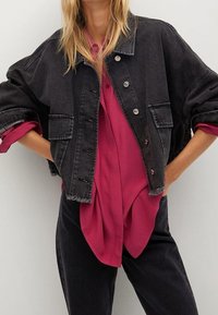 Schwarze Jeansjacke mit fallenden Schultern, zwei Brusttaschen und Metallknöpfen, getragen über einer lockeren, tiefrosa Bluse.