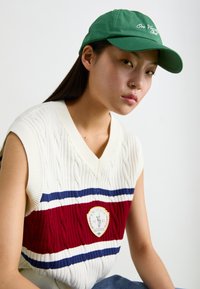 Zelena kapa z vezenim napisom, smetana pulover brez rokavov z rdečimi in modrimi črtami ter osrednjim logotipom, ki ima pleteno teksturo.