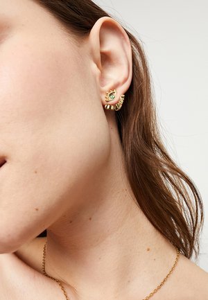 Pendientes de oro con un diseño texturizado de onda y una pequeña gema turquesa, sobre piel bronceada y cabello castaño.