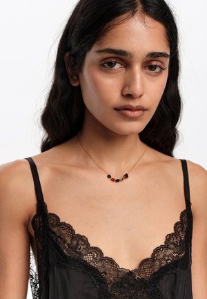 Młoda kobieta z długimi ciemnymi włosami nosi czarny top z koronkowymi wykończeniami oraz złoty naszyjnik z wielokolorowymi kamieniami szlachetnymi na białym tle.