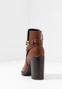Hnědý kotníkový boty z umělé kůže s vysokým blokovým podpatkem, přezkou jako ozdobou a texturovanou podrážkou, s hladkým povrchem a elegantním tvarem.