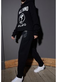 Schwarze, kurze Kapuzenjacke mit weißem "Moschino Milano"-Druck, kombiniert mit schwarzen Sweatpants, die ein Strassbärenmotiv aufweisen. Weiße Sneaker.