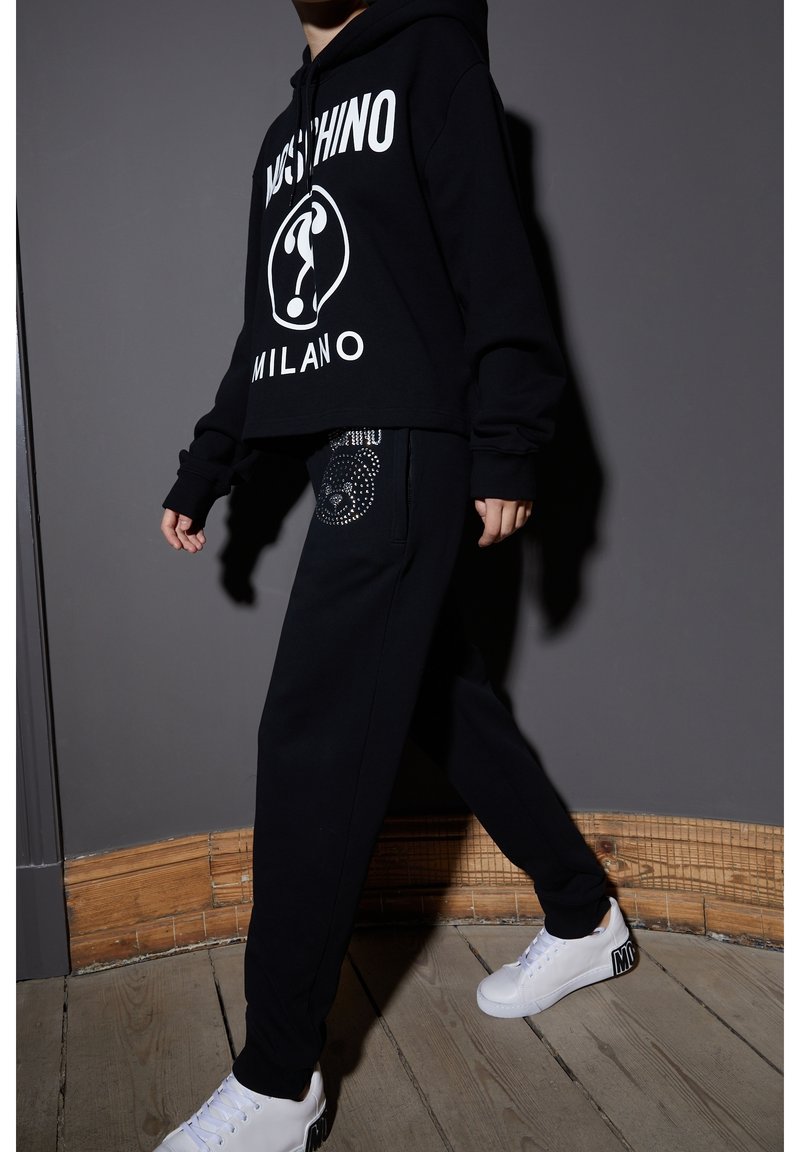 Schwarze, kurze Kapuzenjacke mit weißem "Moschino Milano"-Druck, kombiniert mit schwarzen Sweatpants, die ein Strassbärenmotiv aufweisen. Weiße Sneaker.