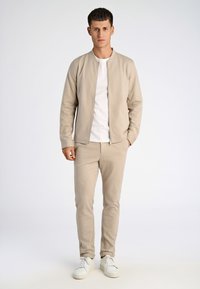 Beige zip-up jacka med en slät textur, i kombination med matchande byxor. Bärs över en vit t-shirt och kompletteras av vita sneakers.