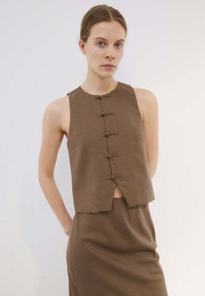 Giovane donna che indossa un top marrone senza maniche con quattro chiusure a bottoni annodati, abbinato a una gonna marrone coordinata, in piedi su uno sfondo semplice.