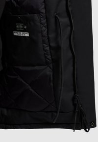 Svart jackas insida med vadderad textur, utrustad med en blixtlåsförsedd ficka och en etikett. Mjuk yttertyg med minimal detaljering.