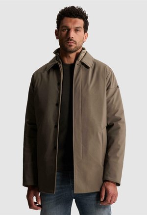 Homme aux cheveux courts et bouclés portant une veste taupe boutonnée sur une chemise noire et un jean bleu, debout devant un fond clair uni.