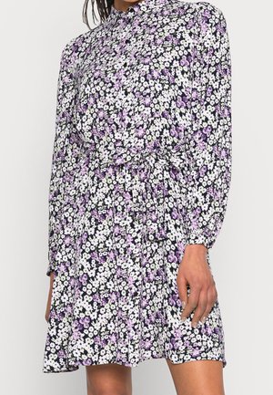 Robe à fleurs à manches longues avec des fleurs blanches et violettes, boutonnée à l'avant, col et ceinture à la taille portée par une personne à la peau de ton moyen-foncé.