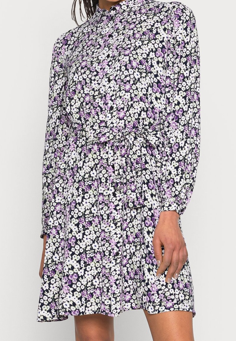 Robe à fleurs à manches longues avec des fleurs blanches et violettes, boutonnée à l'avant, col et ceinture à la taille portée par une personne à la peau de ton moyen-foncé.