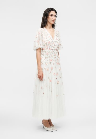 Γυναίκα στέκεται φορώντας λευκό, μακρύ μέχρι τον αστράγαλο φόρεμα με διάφανα φουσκωτά μανίκια και ροζ κεντημένα λουλούδια, σε συνδυασμό με λευκά μυτερά τακούνια.