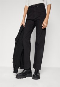 Pantalones negros de cintura alta de mezclilla, con corte recto y diseño clásico de cinco bolsillos, combinados con botines negros y una chaqueta.