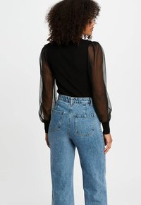 Frau mit lockigem Haar, die ein schwarzes Oberteil mit durchsichtigen Puffärmeln und hochtaillierte blaue Jeans trägt, mit dem Rücken zu einer weißen Wand gewandt.