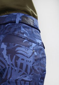 Blå byxor med camouflage-mönster i texturerad tyg, med en läderlapp på midjan och en metallknapp på bakfickan.