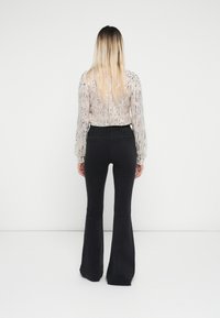 Blouse en voile rose clair avec des motifs texturés et des manches longues, associée à un jean noir évasé. La tenue est vue de dos.