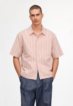 Homme portant une chemise à manches courtes rayée rose clair boutonnée et un pantalon bleu foncé ample, les mains dans les poches.