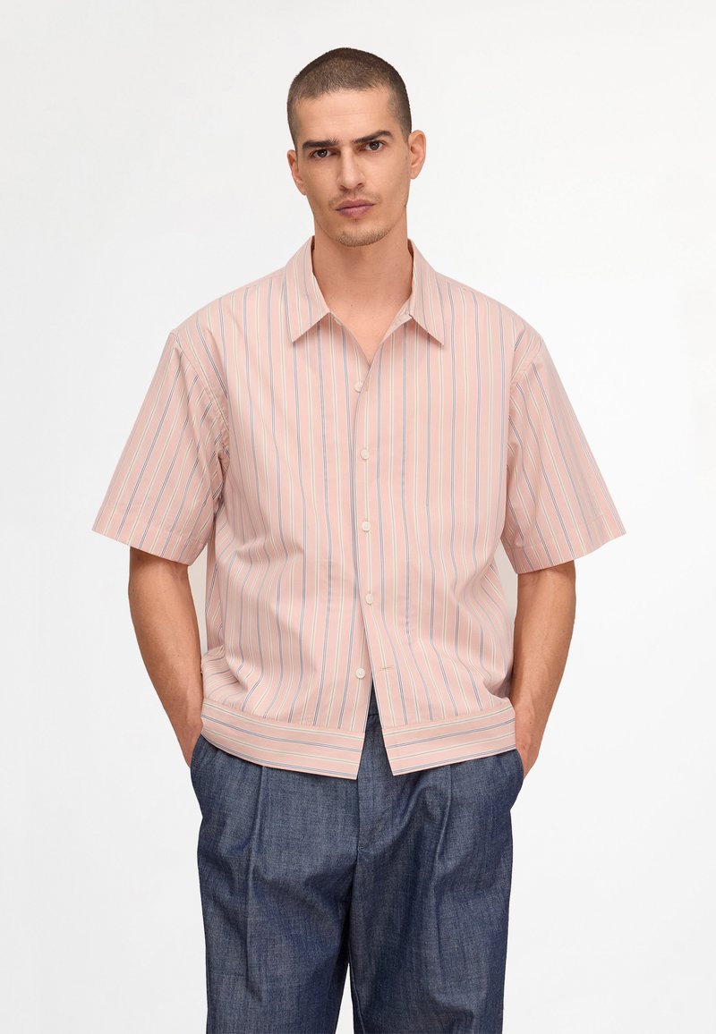 Homme portant une chemise à manches courtes rayée rose clair boutonnée et un pantalon bleu foncé ample, les mains dans les poches.