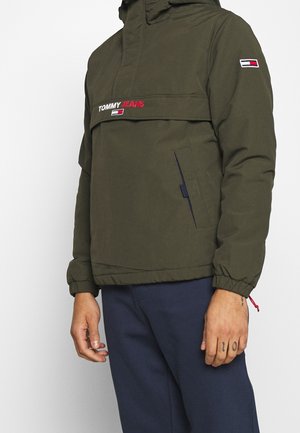 Homme portant une veste Tommy Jeans vert olive avec un logo sur le devant et un pantalon bleu marine, debout devant un fond blanc uni.