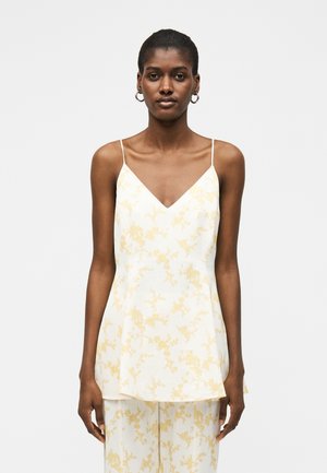 Nő fehér és halványsárga virágmintás ujjatlan topban, vékony pántokkal és hozzáillő nadrággal, egyszínű világos háttér előtt.