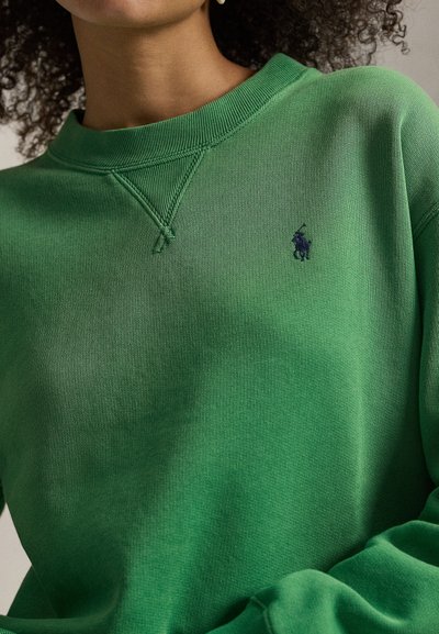 Άτομο που φοράει πράσινο φούτερ με στρογγυλή λαιμόκοψη και σκοτεινό κεντημένο λογότυπο παίκτη πόλο στο αριστερό στήθος, με εμφανή σγουρά μαλλιά.