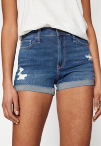Mørkeblå denimshorts med en frynsedekorasjon nederst, slitte detaljer foran, og en brettet kant i bunnen. Standard fem-lommersdesign.