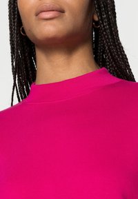 Sweatshirt fuchsia avec col montant, texture côtelée et manches courtes, avec des détails de couture autour de l'encolure.