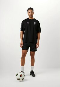 Μαύρο βαμβακερό μπλουζάκι με κοντά μανίκια και σορτς, διακοσμημένα με λογότυπα. Το μοντέλο στέκεται με μια μαύρη ποδοσφαιρική μπάλα σε απλό φόντο.