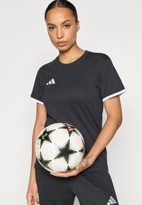 Μαύρο αθλητικό μπλουζάκι με κοντά μανίκια και λευκές λεπτομέρειες, σε συνδυασμό με μια μαύρη και λευκή ποδοσφαιρική μπάλα με σχέδια αστεριών και πολύχρωμες λεπτομέρειες.