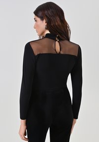 Donna con capelli lunghi e scuri che indossa una maglia nera a maniche lunghe con un pannello trasparente sulla parte superiore della schiena e un dettaglio a occhiello con bottoni sul collo.