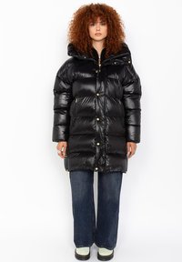 Sort puffers jakke med en skinnende tekstur, oversized pasform, hætte og gyldne hardware-detaljer. Parret med vide jeans og lyse støvler.