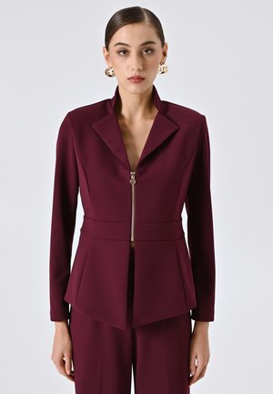 Frau in einem tief burgunderfarbenen Blazer mit Reißverschluss und Spitzkragen, kombiniert mit maßgeschneiderten Hosen in der gleichen Farbe, accessoriert mit goldenen Kreolen.