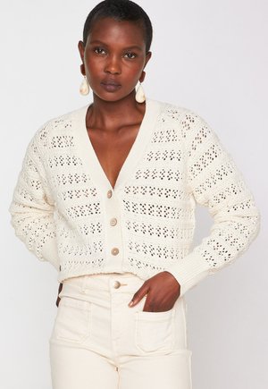 Cardigan all'uncinetto in crema con scollo a V, chiusura con bottoni, polsini a coste e motivo a maglia aperta. Abbinato a jeans chiari.