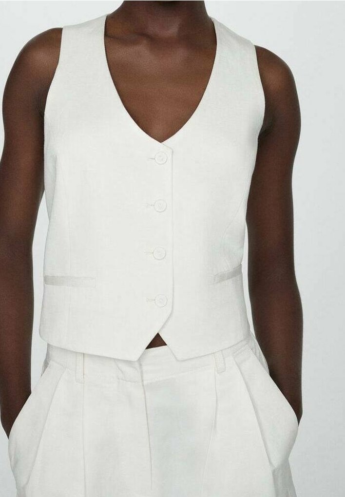 Personne portant un gilet blanc ajusté sans manches boutonné et un pantalon taille haute assorti avec les mains dans les poches.