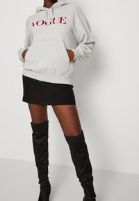 Felpa grigia con "VOGUE" in lettere rosse, tasca frontale, abbinata a una mini gonna nera e stivali neri sopra il ginocchio.