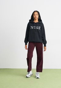 Crna kapuljača s "NIKE" logotipom, bordo kord ušivene široke hlače i bijele tenisice. Jednostavna pozadina s zelenim podom.