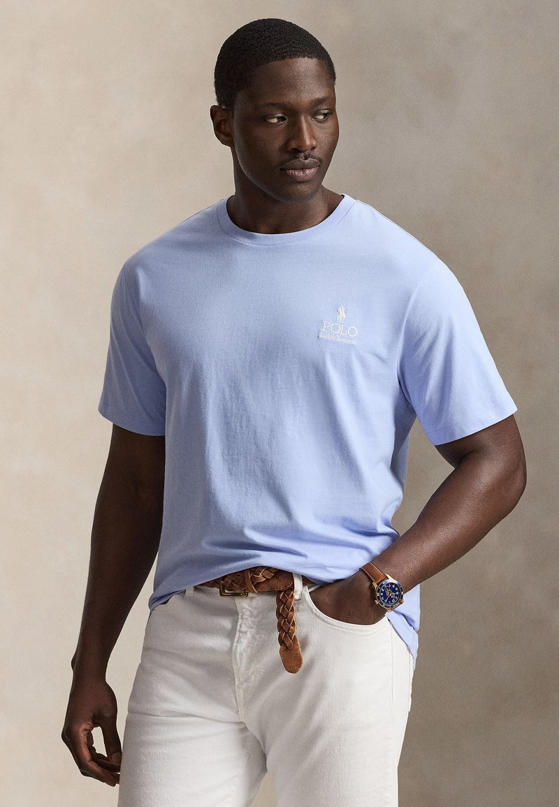 Άνδρας που φοράει ανοιχτό μπλε μπλουζάκι Polo Ralph Lauren, λευκό παντελόνι, καφέ πλεγτή ζώνη και μπλε ρολόι χειρός, κοιτάζει προς τα αριστερά.
