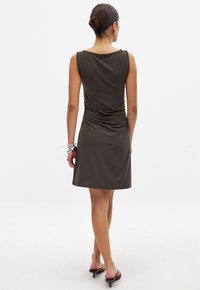 Robe sans manches de couleur brun foncé avec une silhouette ajustée, des détails froncés à l'arrière et un tissu lisse et doux. Portée avec des sandales simples.