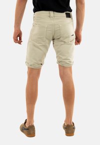 Lichtbeige shorts met opgerolde omslagen, achterzakken en een leren patch op de tailleband. Draagt casual bruine schoenen.