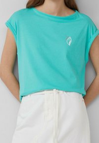 Turkos kortärmad topp med uppvikta ärmar och ett litet vitt snäckskalsmärke, ihop med högmidjade vita byxor med bälteshällor.