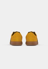 Um par de sapatos casuais amarelo mostarda com solas castanhas, vistos de trás, sobre um fundo branco liso.