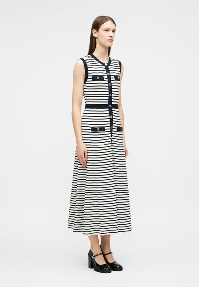 Jeune femme portant une robe midi sans manches à rayures noires et blanches avec des boutons et des escarpins Mary Jane noirs, debout devant un fond blanc.