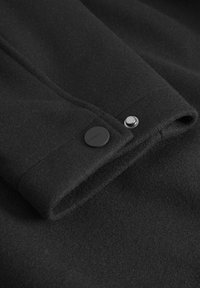 Črna volnena tkanina z gladko teksturo, ki vsebuje detajl gumba in subtilno šivanje ob robovih. Kompaktna in strukturirana zasnova.