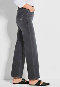 Graue Jeans im Straight-Leg-Schnitt mit Seitentaschen und dekorativer Stickerei auf den Gesäßtaschen. Getragen mit schwarzen Slippern.