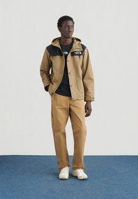 Καφέ αδιάβροχο μπουφάν με μαύρο πάνω μέρος, σετ με καφέ παντελόνια και λευκά αθλητικά παπούτσια. Το μπουφάν διαθέτει κουκούλα και κλείσιμο με κουμπιά.